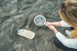 Eine junge Frau hockt am Strand und filtert mit einem Sieb Plastikpartikel aus dem Wasser.