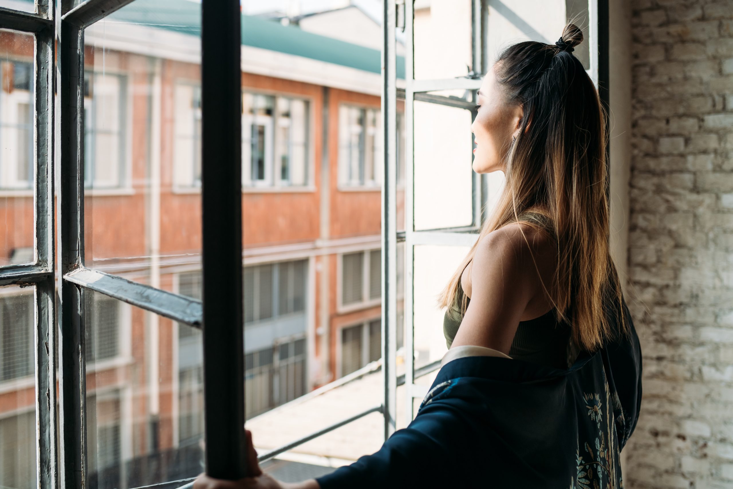 Eine junge Frau steht an einem zum Lüften geöffneten Fenster und sieht nach draußen.